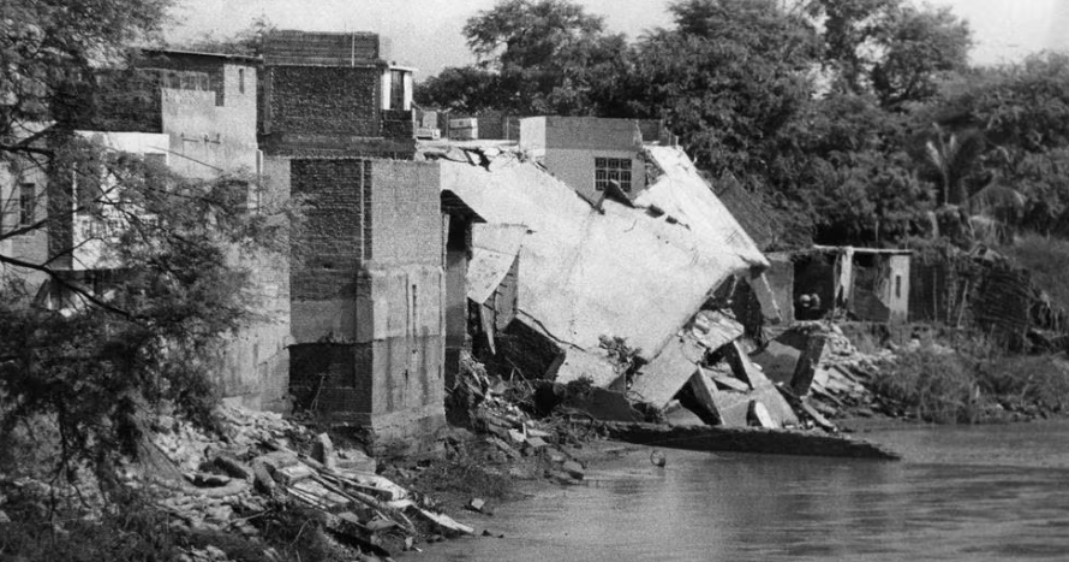 Fotografía en blanco y negro de un edificio colapsado con escombros cayendo sobre la orilla de un río.